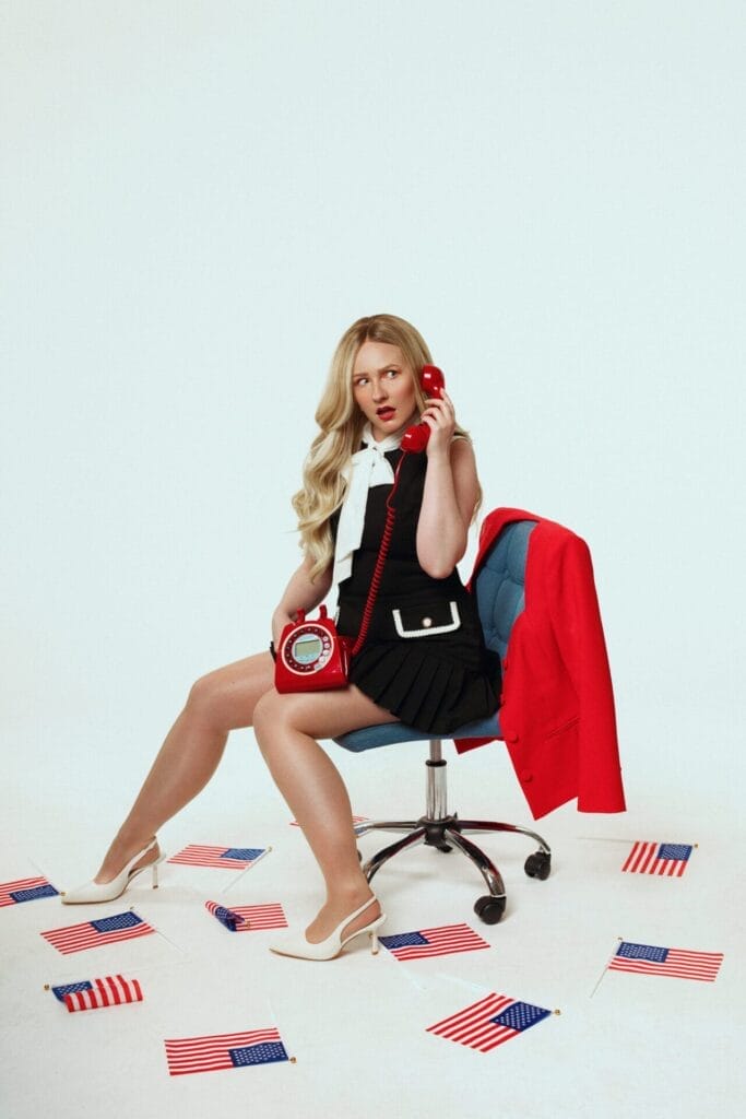 Leigh Douglas with long blonde hair over one shoulder, sitting on a swivel chair, wearing a black dress, a white scarf, and white kitten heels, holding a red dial phone to her ear with the dial on her lap. The floor is scattered with hand-waving USA flags, and there's a red jacket on the back of the chair,