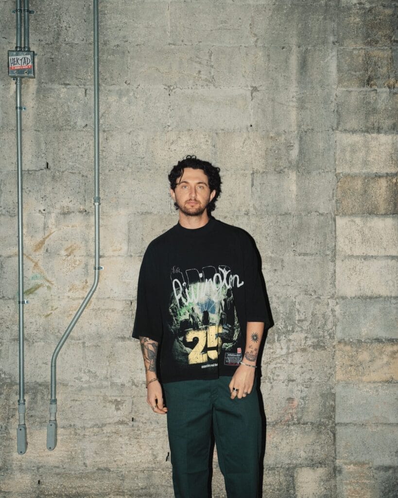 Maz Styler with tattoos on his arms stands against a textured concrete wall, wearing a black T-shirt with "25" printed on it, and green pants. The mood is casual and urban.