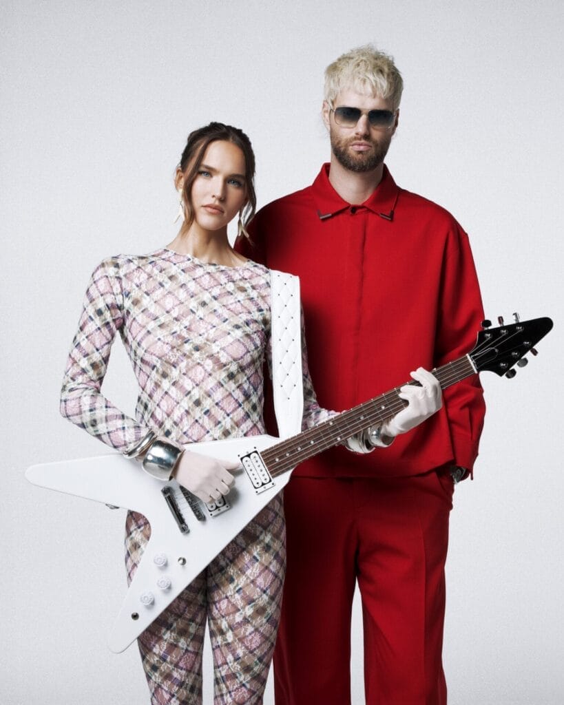 SOFI TUKKER Launch New Chapter With J Balvin Collaboration “Cook” and Republic Records Signing 46 SOFI TUKKER posing together where Sophie Hawley-Weld is dressed in a patterned jumpsuit and Tucker Halpern is in a red outfit standing confidently. She holds a white electric guitar, conveying a modern, stylish look.