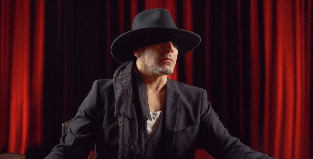 Ray Vale in a black suit and wide-brimmed hat and a black eye mask sits against a backdrop of red curtains, exuding a mysterious and sophisticated vibe.