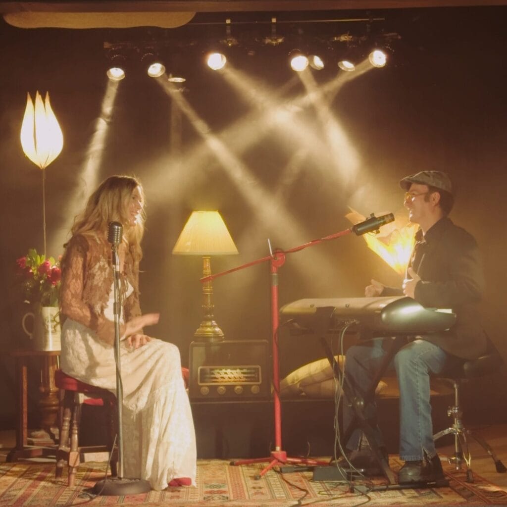 Joss Stone in a lace dress sings into a vintage microphone beside Conner Reeves playing a keyboard. They're illuminated by warm stage lights, creating an intimate, retro feel.