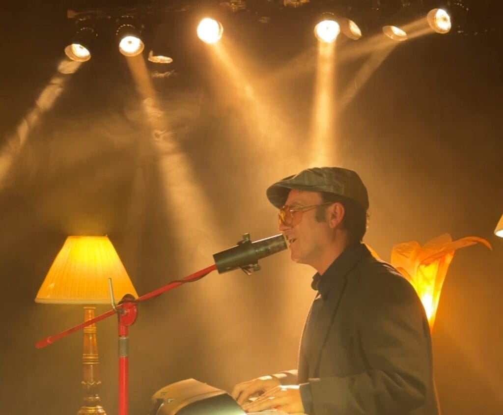 Conner Reeves wearing a flat cap and glasses plays a keyboard under warm stage lights. A vintage lamp and soft yellow lighting create an intimate, cosy ambiance.