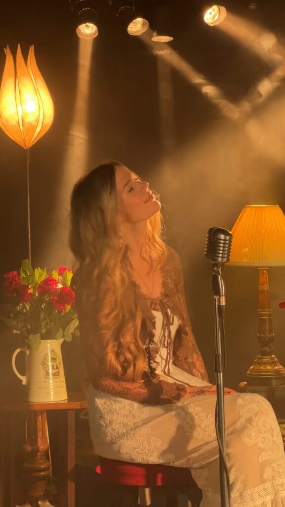 Joss Stone sits on a stool on stage in a dimly lit room, her eyes closed and head tilted slightly back. Vintage mic, soft yellow lamps, a vase with red roses nearby.