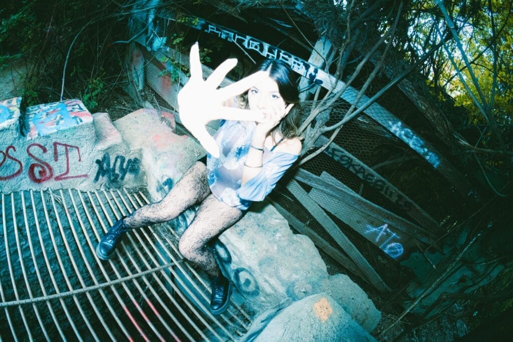 Hailey Hermida sits on graffiti-covered concrete steps, reaching toward the camera. The scene has an edgy, artistic feel with surrounding foliage.
