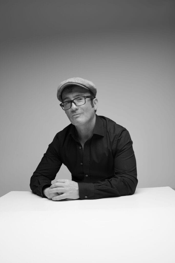 Black and white photo of Conner Reeves in a flat cap and glasses, sitting at a table with hands clasped, exuding a confident and relaxed demeanour.