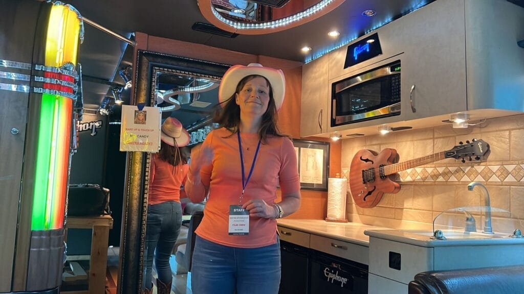 Donna Crowe in a white cowboy hat and orange shirt stands inside a tour bus kitchen. Neon lights and a guitar on the wall create a cosy, musical ambiance.