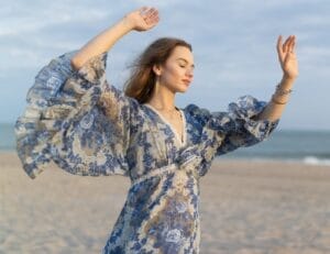 Lina Maxine in a flowing, blue-patterned dress dances gracefully on a beach, her eyes closed and arms raised, conveying a sense of freedom and serenity.