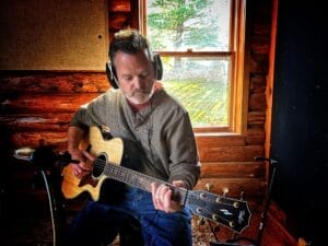 Fred Presley with headphones plays an acoustic guitar in a rustic wooden room, focused and calm. Sunlight streams through a window, creating a warm ambiance.
