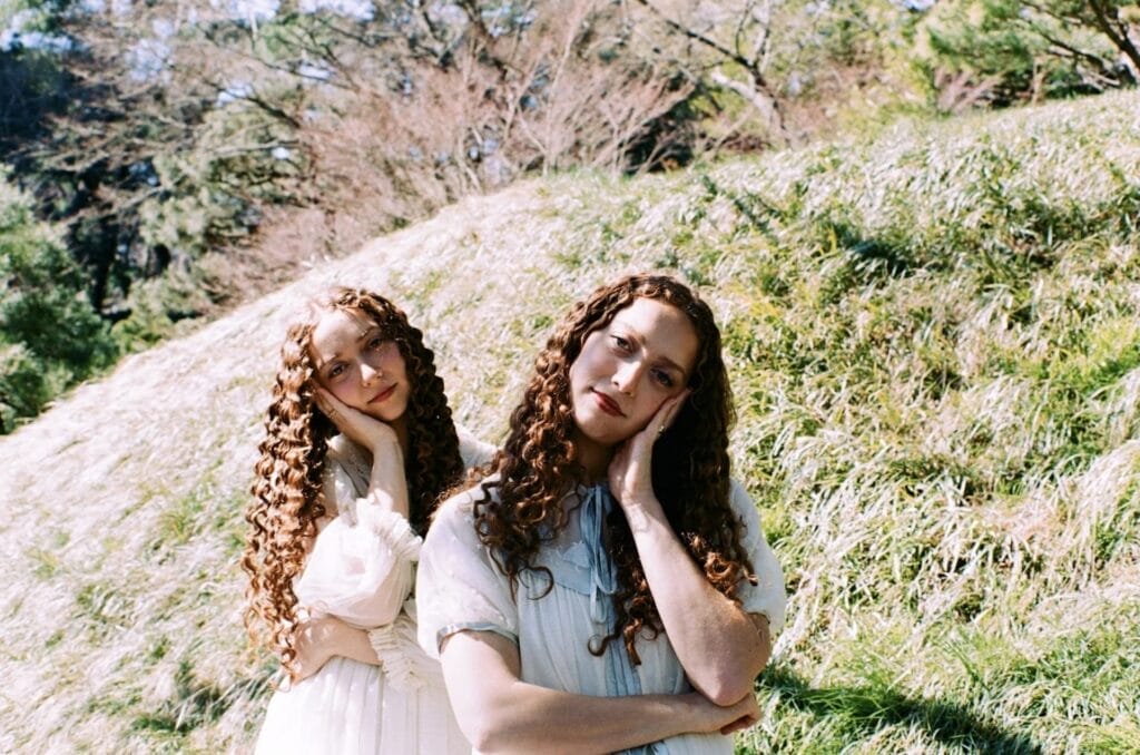 Ruby & Sasha with long curly hair are standing on a grassy hillside, their heads tilted and hands resting on their cheeks. The scene is serene and natural.