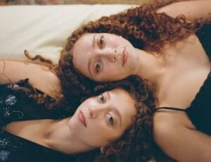 Twins Ruby & Sasha with curly hair lie side by side, gazing softly at the camera. They wear dark clothing. The tone is calm and intimate.