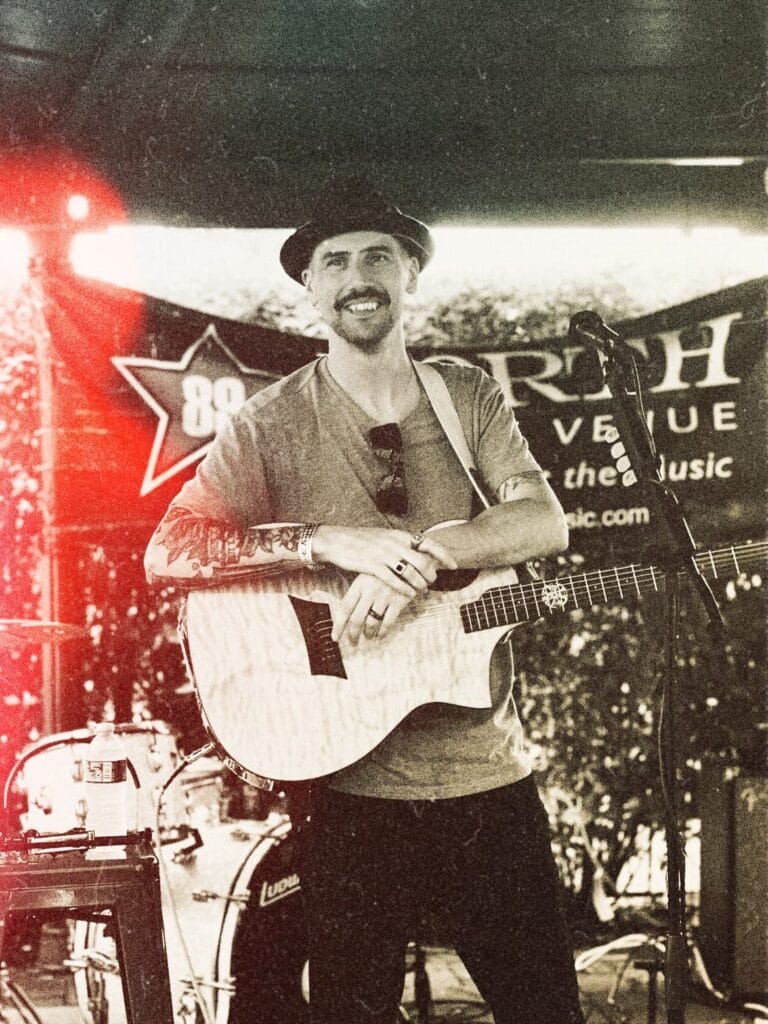 Nicholas Lopardo as Minimum Champion, smiling on stage with an acoustic guitar, wearing a hat. The background features band equipment and a banner, giving a warm, vintage vibe.