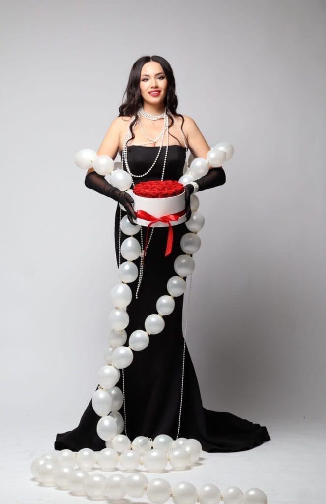 Olena Grytsynger dressed in a long black dress and gloves holds a white box with red roses, adorned with a red ribbon. She is surrounded by pearl-like balloons, creating an elegant and celebratory atmosphere.