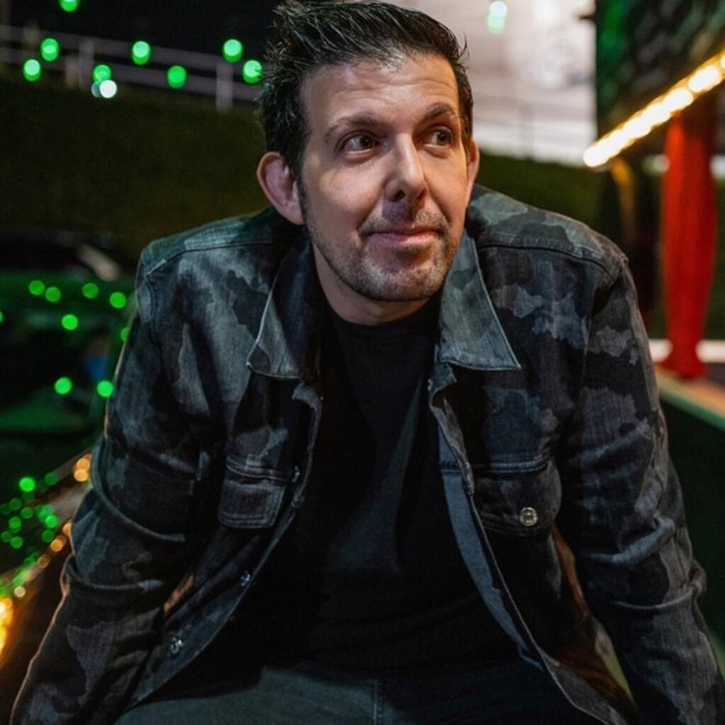 Jon Sosis with short dark hair, wearing a camo-patterned jacket, is sitting outdoors at night. Green lights in the background suggest a lively atmosphere.