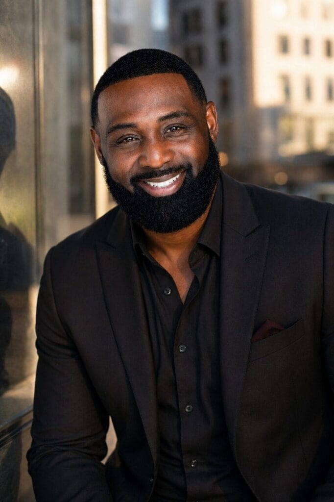 Sean Anthony Baker in a black suit sits outdoors with an urban backdrop. Sunlight highlights his face, creating a warm and confident tone.
