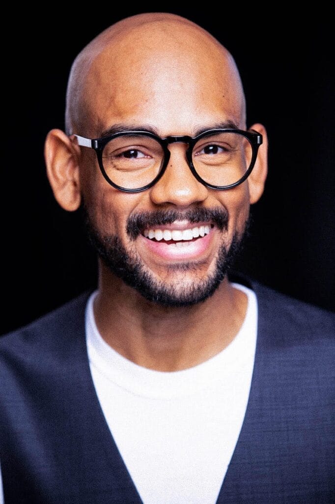 Todd A. Jackson who has a bald head, a beard and wears round glasses, a white shirt, and a dark vest is in front of a black background, conveying warmth and friendliness.